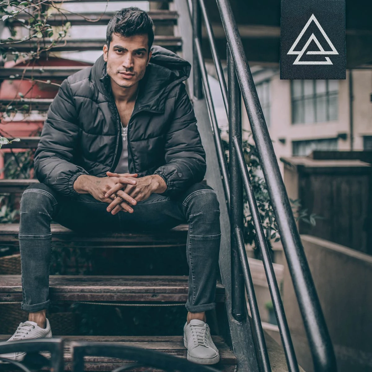 Man in a black puffer jacket sitting on outdoor stairs, with the APODOKIA patch top right