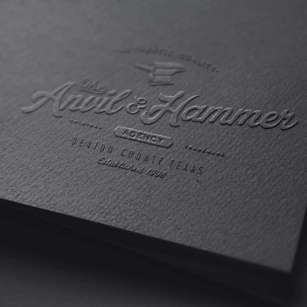 The Anvil & Hammer Agency logo blind-embossed on dark textured paper