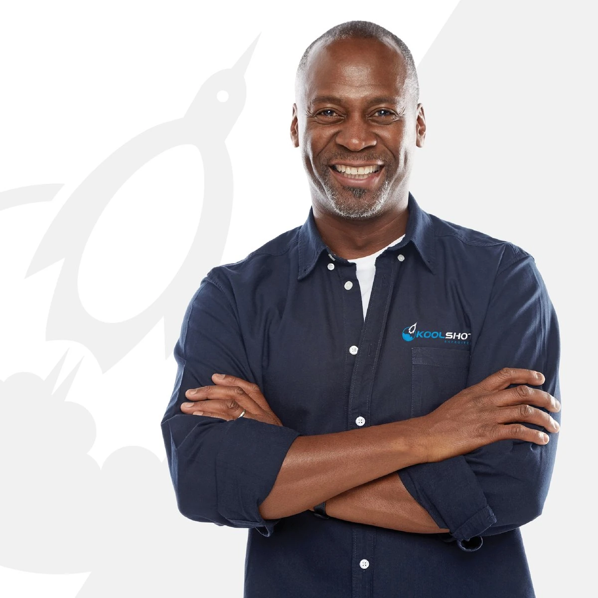 Smiling crew member in a navy KoolShot shirt with arms crossed, in front of a large faint penguin mark