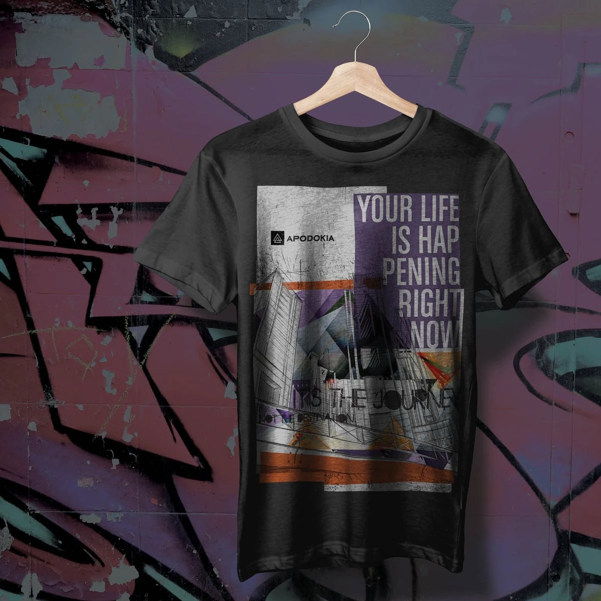 Black APODOKIA graphic tee reading “Your life is happening right now” on a hanger against a graffiti wall