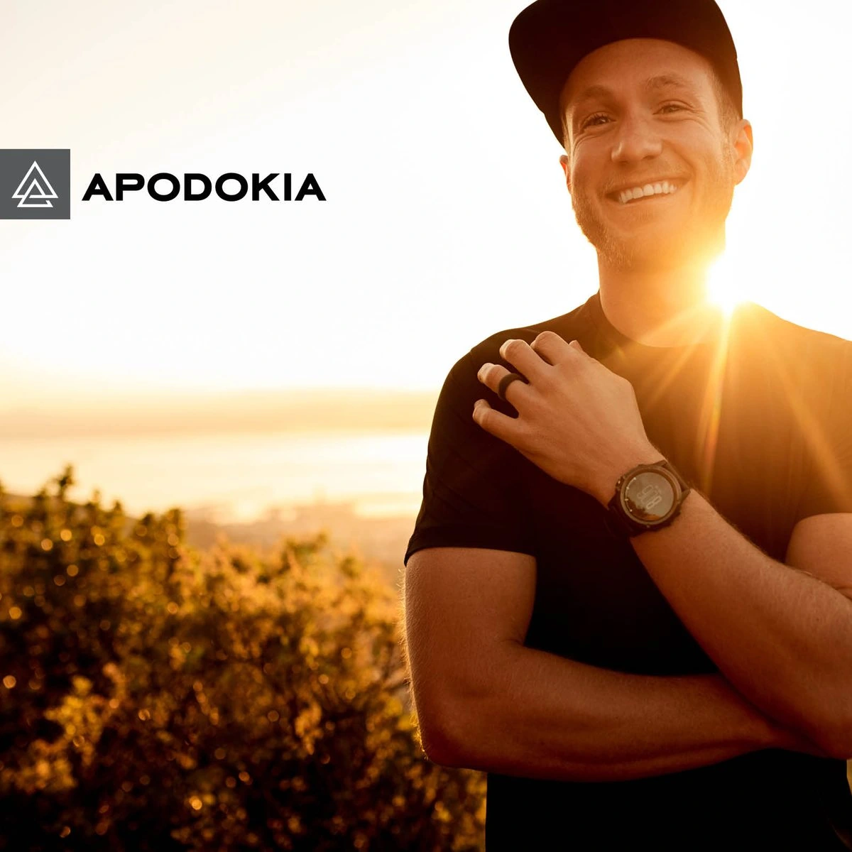 Smiling man in a cap at sunset with his hand on his chest, next to the APODOKIA logo