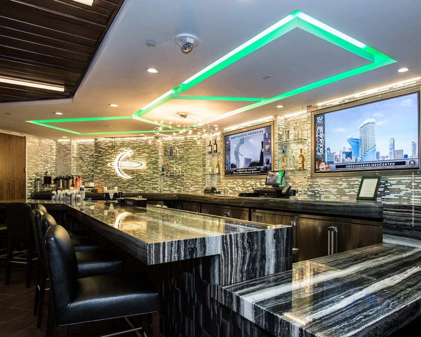 Suite-level bar with green ceiling lighting, a marble counter and screens showing American Airlines Center content