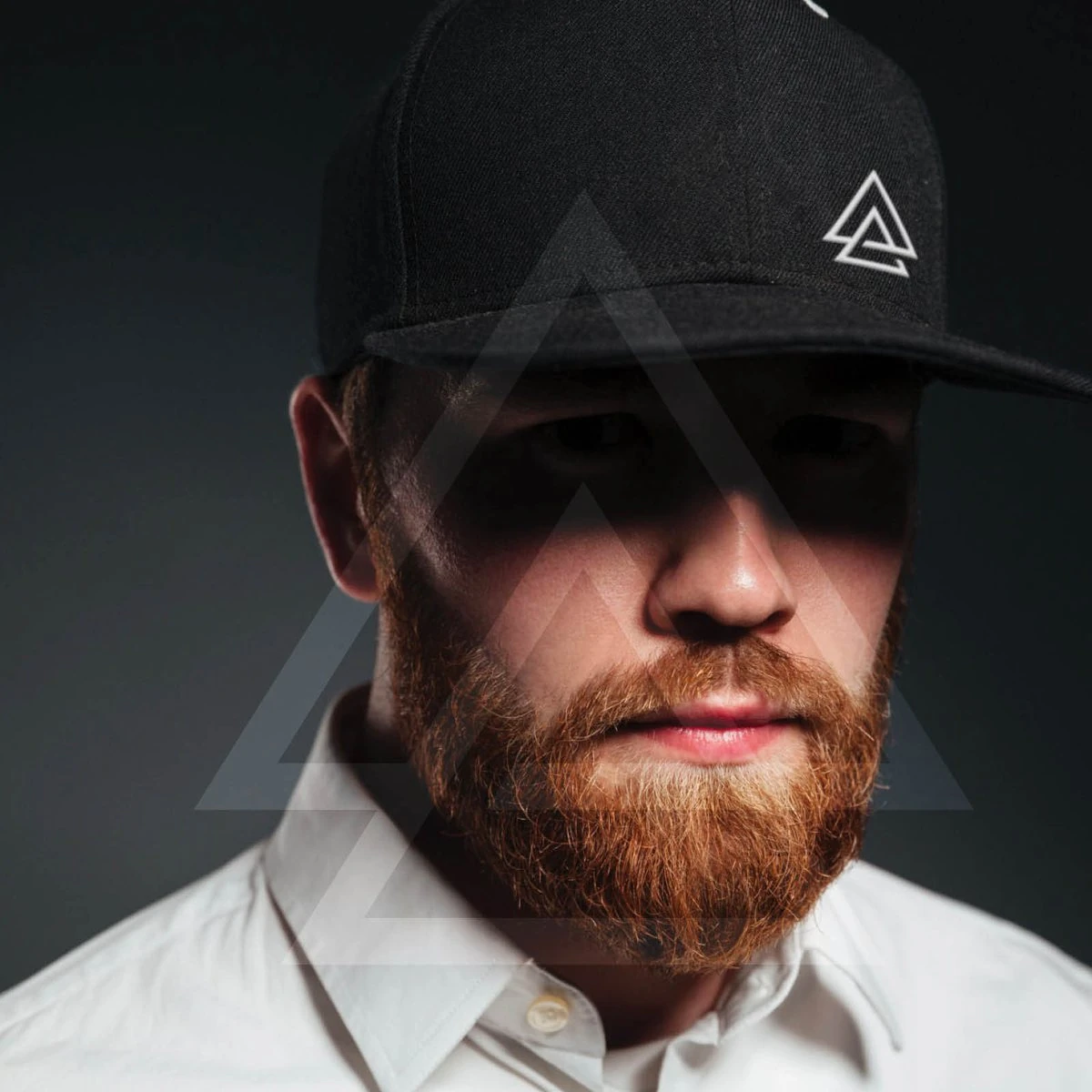 Bearded man in a black snapback with the APODOKIA mark, large faint triangles over the portrait