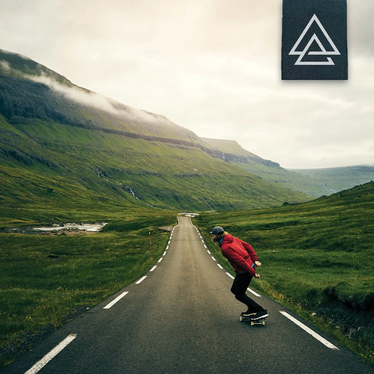 A skateboarder in a red jacket rides down an empty valley road, with the APODOKIA patch in the corner