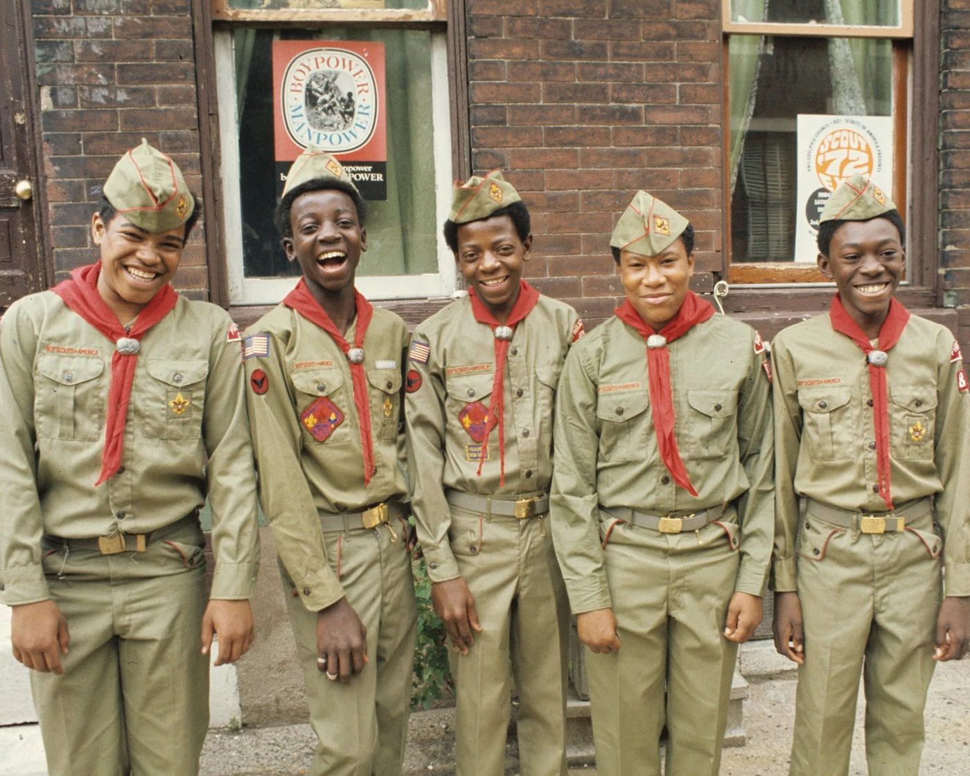 Five smiling Scouts in 1970s uniforms and red neckerchiefs in front of a brick building