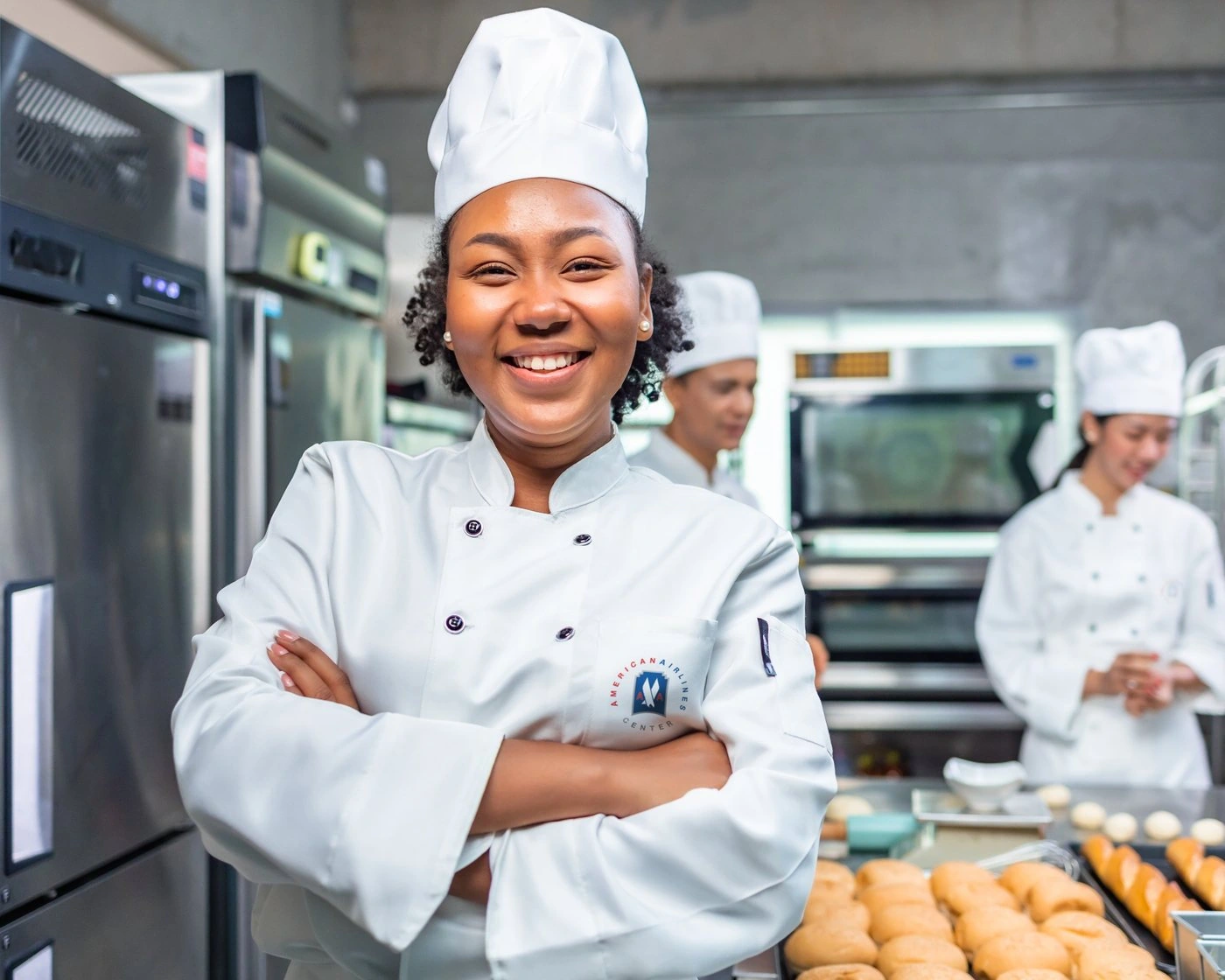 Smiling chef in a white jacket with the American Airlines Center logo, arms crossed in a bakery kitchen